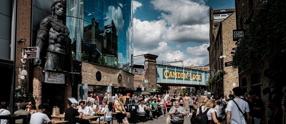 A busy street scene in Fitzrovia, London, showing numerous pedestrians walking and sitting at outdoor tables of cafes and restaurants. The area features a mix of modern glass-fronted buildings and tra