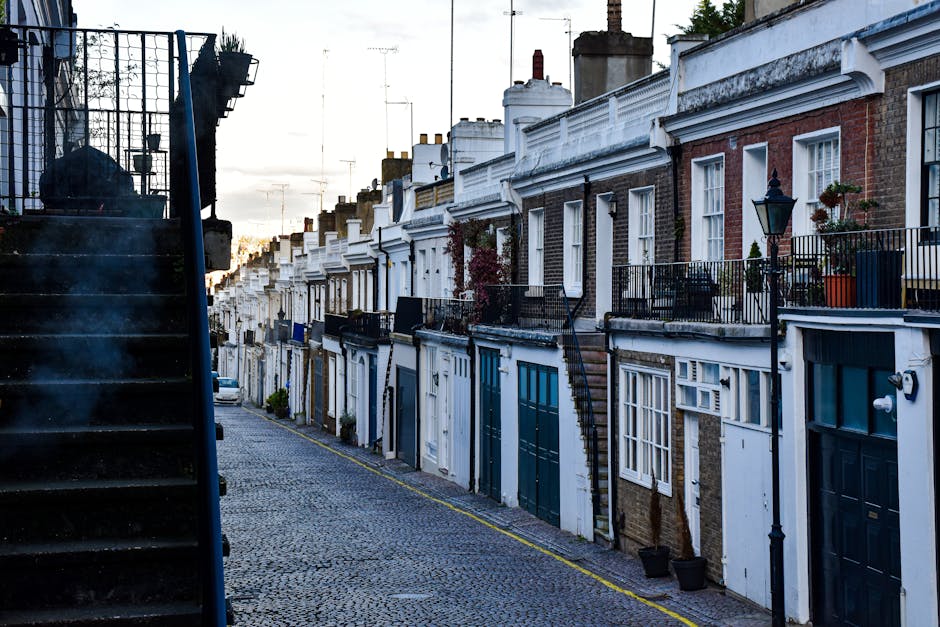 A view of a narrow residential street in Fitzrovia during daylight, showing a row of terraced houses with brick facades, white decorative moldings, and small balconies with black metal railings. Sever