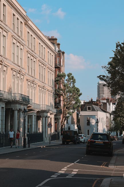 The image shows a quiet residential street in Fitzrovia with a row of elegant, multi-storey Victorian-style buildings featuring large windows, ornate balconies, and decorative moldings, illuminated by natural daylight. The street is lined with several parked cars, including a black van partially visible near the curb, and a few pedestrians walking along the pavement. Leafy trees provide greenery along the sidewalk, casting shadows on the buildings and road. In the background, modern high-rise buildings contrast with the historic architecture, under a partly cloudy sky. The scene depicts a typical urban setting suitable for home relocation and furniture transport, with visible elements such as parked vehicles, building facades, and street furniture supporting the logistics of packing and moving services carried out by companies like Man and Van Fitzrovia.