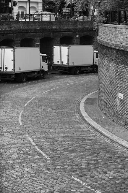 A busy city street scene in Fitzrovia featuring a historic multi-storey building with ornate architectural details, balconies, and a pointed roof. Parked vehicles include a white moving van branded with 'manandvanfitzrovia.co.uk', a yellow taxi, and several motorbikes. The loading area is on the pavement adjacent to the building, with some equipment such as straps and blankets likely used for furniture transport visible nearby. Street poles, traffic lights, and road markings are present to support safe loading and unloading processes during home relocation. In the background, modern skyscrapers and a clear blue sky with some wispy clouds complete the urban setting. The scene captures the typical logistics of packing and moving services in a central London neighbourhood, emphasizing the importance of careful furniture transport and efficient removal strategies as seen by Man and Van Fitzrovia.