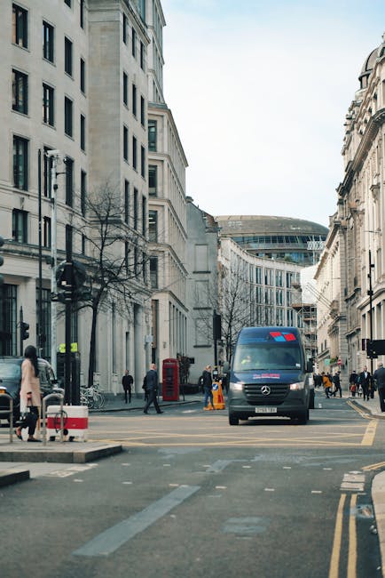 A city street scene showing a blue Mercedes-Benz van parked at a loading zone during daytime, with several pedestrians walking on the sidewalk and crossing the road. The surrounding environment features tall, light-colored stone and concrete buildings with multiple windows, some with decorative architectural elements, and leafless trees indicating a winter season. The van is positioned near the entrance to a building, and orange traffic cones are placed on the pavement nearby. In the background, a modern curved glass building is visible, and the sky is partly cloudy with natural daylight illuminating the area. This setting illustrates a typical urban home relocation process involving furniture transport and loading activities, relevant to the services provided by Man and Van Fitzrovia for efficient packing and moving logistics.