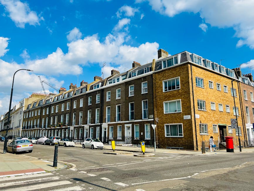 A row of multi-story residential buildings with brick facades and multiple windows lines a busy street in Fitzrovia. Some windows are open, revealing interior curtains and furnishings, while others are closed. The street features parked cars, a lamppost, and road signage, with a red post box near the pavement. The sky is bright blue with scattered white clouds, and a construction crane is visible in the background. Man and Van Fitzrovia, a professional removals company, often transports household furniture and boxes during home relocation processes, which may involve loading and unloading items in similar urban environments. Inside the property, moving blankets, cardboard boxes, and furniture can be seen, indicating packing and preparing for furniture transport as part of a house removal service.