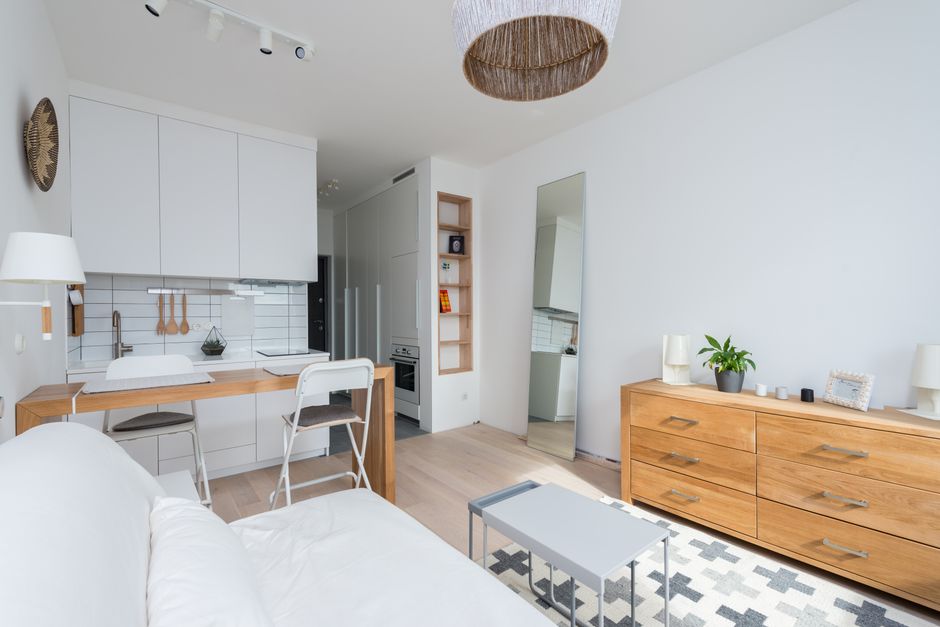 Interior view of a modern, minimalist studio flat with white walls and light wood flooring, showcasing a compact kitchen area with white cabinetry, a stainless steel sink, and a white tile backsplash. Two white chairs are positioned at a small wooden breakfast bar, which is adjacent to the kitchen. A white sofa with a loose cushion is visible in the foreground, partially out of frame. To the right, a wooden sideboard with a natural finish holds small decorative items, a potted plant, and table lamps. A tall, freestanding mirror leans against the wall near the kitchen, reflecting part of the room. The room is well-lit with natural light, and a fabric-covered ceiling light fixture hangs overhead. This scene depicts a typical home environment preparing for a house relocation, with a focus on the interior layout and furnishings suited for a moving process, as managed by a house removals specialist like Man and Van Fitzrovia.