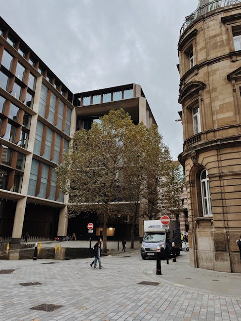 A street scene in Fitzrovia showing a modern multi-story office building with large glass windows and a concrete facade on the left and a historic curved stone building with detailed architecture on the right. In the foreground, a tree with sparse leaves extends over the paved pedestrian area, which features several bollards and manhole covers. Two delivery vans are parked nearby, one with visible branding, and a few people are walking across the street, some wearing casual clothing. The sky is overcast, creating diffuse lighting. The context suggests a busy urban environment suitable for house removals and furniture transport services, with potential logistical activity related to moving and loading in the area, aligning with Fitzrovia same day removals near Fitzroy Square as listed on manandvanfitzrovia.co.uk.