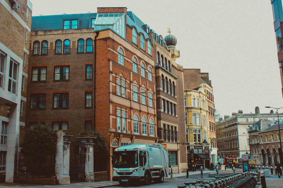 The image depicts a street view in Fitzrovia, featuring a light blue removal truck parked outside a multi-storey brick and stone building with large windows, which is likely being used for home relocation purposes. The truck, associated with a professional removals service, is positioned near the building's entrance on the pavement, with visible loading equipment, including a trolley and wrapped furniture. Adjacent to the truck, there are several cardboard boxes and blankets used for packing and protecting furniture during the moving process. The street is lined with other historic and modern buildings, with some in yellow and beige tones, and there are bicycles parked along the roadside. Street lamps and bollards are present, indicating an urban environment suitable for furniture transport and moving logistics. This scene captures the typical loading process involved in furniture transport and packing and moving services provided by companies like Man and Van Fitzrovia, reflecting the careful handling of items during a house removal or relocation in the Fitzroy Square area.