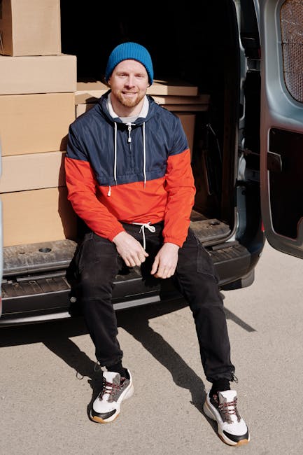 A man with a beard, wearing a blue knit beanie, a navy blue and red waterproof jacket, black trousers, and white sneakers, is sitting on the edge of an open loading area of a grey van parked on a street. He is surrounded by stacked cardboard boxes of various sizes, some with packing tape and others with plastic wrap. The van’s interior is visible, showing more boxes and packaging materials, indicating an ongoing home relocation or furniture transport process. The man appears to be part of a professional removals service, possibly assisting with packing and moving tasks for a small flat in Fitzrovia. The loading area is illuminated by natural daylight, and the pavement is visible beneath his feet, reflecting the typical environment for household moves carried out by Man and Van Fitzrovia.