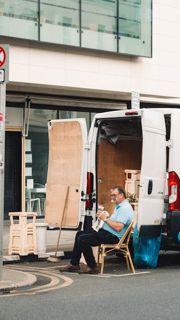 A man wearing glasses, a light blue shirt, and dark trousers is seated on a wooden chair just outside the open rear doors of a white cargo van parked on a city street. The van is loaded with packed cardboard boxes, wooden panels, and protective packaging materials, indicating a home relocation or furniture transport process. The man is using a mobile device, possibly coordinating or taking a break during packing and moving activities. In the background, there is a modern building with large glass windows and a balcony. The van is positioned next to the pavement, with some plastic-wrapped furniture and additional packing materials nearby. This scene reflects the logistics chain involved in a small flat or house move, typical of services provided by Man and Van Fitzrovia, supporting efficient house removals and moving services.