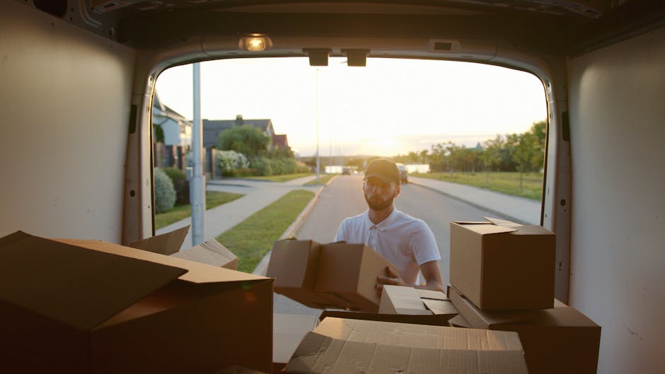 A man with a beard, wearing a blue knit beanie, a navy blue and red waterproof jacket, black trousers, and white sneakers, is sitting on the edge of an open loading area of a grey van parked on a street. He is surrounded by stacked cardboard boxes of various sizes, some with packing tape and others with plastic wrap. The van’s interior is visible, showing more boxes and packaging materials, indicating an ongoing home relocation or furniture transport process. The man appears to be part of a professional removals service, possibly assisting with packing and moving tasks for a small flat in Fitzrovia. The loading area is illuminated by natural daylight, and the pavement is visible beneath his feet, reflecting the typical environment for household moves carried out by Man and Van Fitzrovia.