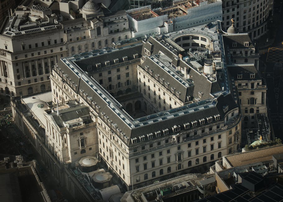 An aerial view of a historic, multi-storey office building with a central courtyard, situated within a dense urban area in Fitzrovia, London. The building features a classic architectural style with a flat roof, extensive rows of windows, and decorative stonework along the façade. Surrounding buildings include other commercial and office properties with similar architectural details. The street level shows narrow roads with parked vehicles and minimal pedestrian activity. The scene is illuminated by natural daylight, highlighting the building's exterior materials and urban environment. This image relates to house and office removals, illustrating the type of property managed by Man and Van Fitzrovia during professional packing and furniture transport services for relocations within central London districts like W1, WC1.
