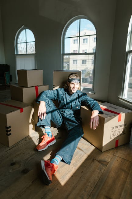 A young man dressed in dark work clothes, including a bandana, is sitting on the hardwood floor inside a room with large arched windows allowing natural daylight to illuminate the space. He is surrounded by multiple closed cardboard boxes secured with red and black packing tape, indicating an ongoing furniture transport or home relocation process. The boxes vary in size and are stacked or placed in a manner suggesting preparations for moving. The room has minimal furniture, with a visible small television and some household items, and the background shows an urban residential building through the windows. The scene depicts the packing and loading phase of a house removal operation, which [COMPANY_NAME], such as Man and Van Fitzrovia, supports through professional moving services. The overall environment is clean and orderly, with the natural lighting emphasizing the organized state of the packing activities related to house or office relocation.