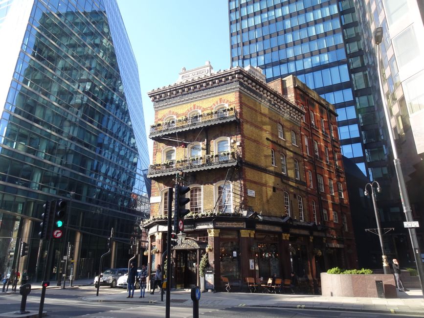 Photograph of a narrow, multi-story brick building situated at a busy street corner in Fitzrovia, with modern glass high-rise buildings on both sides. The older building features ornate iron balconies with decorative railings, large multi-pane sash windows, and a ground-floor commercial space with large glass windows and a black awning. The scene includes traffic lights, pedestrians on the pavement, and several parked cars. The lighting indicates daytime with bright sunlight casting shadows. This setting illustrates the urban environment typical of house and office removals, highlighting the importance of professional furniture transport and packing during moving processes, as seen in the context of small business relocations in Fitzrovia by Man and Van Fitzrovia.