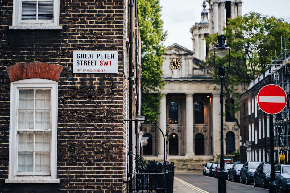 A close-up of the corner of a brick building on Great Peter Street in Westminster, showing a white window with multiple panes and a red brick arch above it. Mounted on the brick wall is a street sign indicating 'Great Peter Street SW1' within the City of Westminster. In the background, a historic building with tall columns and a clock face is visible, along with a black street lamp and a red and white no-entry traffic sign positioned on the street. The scene includes parked cars along the curb and some green trees with foliage, suggesting an urban setting prepared for the logistics of house removals or furniture transport. The lighting suggests daytime, with clear visibility of the environment, walls, and street elements, supporting the context of moving services and logistical planning by Man and Van Fitzrovia.