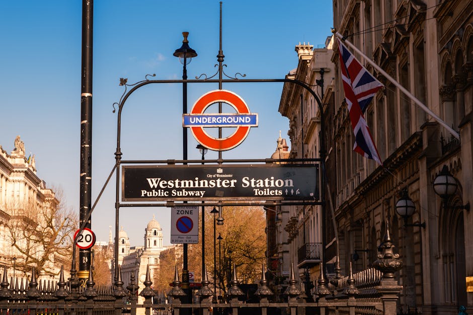 Close-up view of the Westminster Underground station entrance sign on a sunny day, with a Union Jack flag on a pole to the right. The sign indicates the station services the public subway and provides directions to toilets. In the background, historic buildings with intricate architectural details are visible, along with a black lamppost and a metal fence surrounding the station entrance. The scene is typical of central London, with clear blue skies and autumnal trees. This setting reflects an urban environment where a house removal or furniture transport operation by Man and Van Fitzrovia may take place, illustrating the proximity to key transport infrastructure and the typical street scene involved in home relocation and moving logistics.