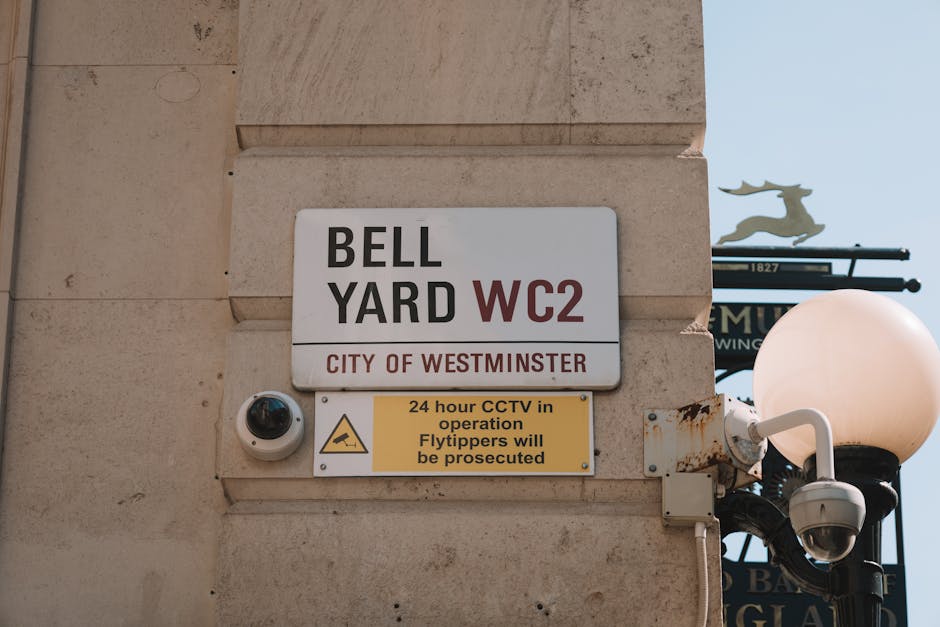 A close-up of the corner of a brick building on Great Peter Street in Westminster, showing a white window with multiple panes and a red brick arch above it. Mounted on the brick wall is a street sign indicating 'Great Peter Street SW1' within the City of Westminster. In the background, a historic building with tall columns and a clock face is visible, along with a black street lamp and a red and white no-entry traffic sign positioned on the street. The scene includes parked cars along the curb and some green trees with foliage, suggesting an urban setting prepared for the logistics of house removals or furniture transport. The lighting suggests daytime, with clear visibility of the environment, walls, and street elements, supporting the context of moving services and logistical planning by Man and Van Fitzrovia.